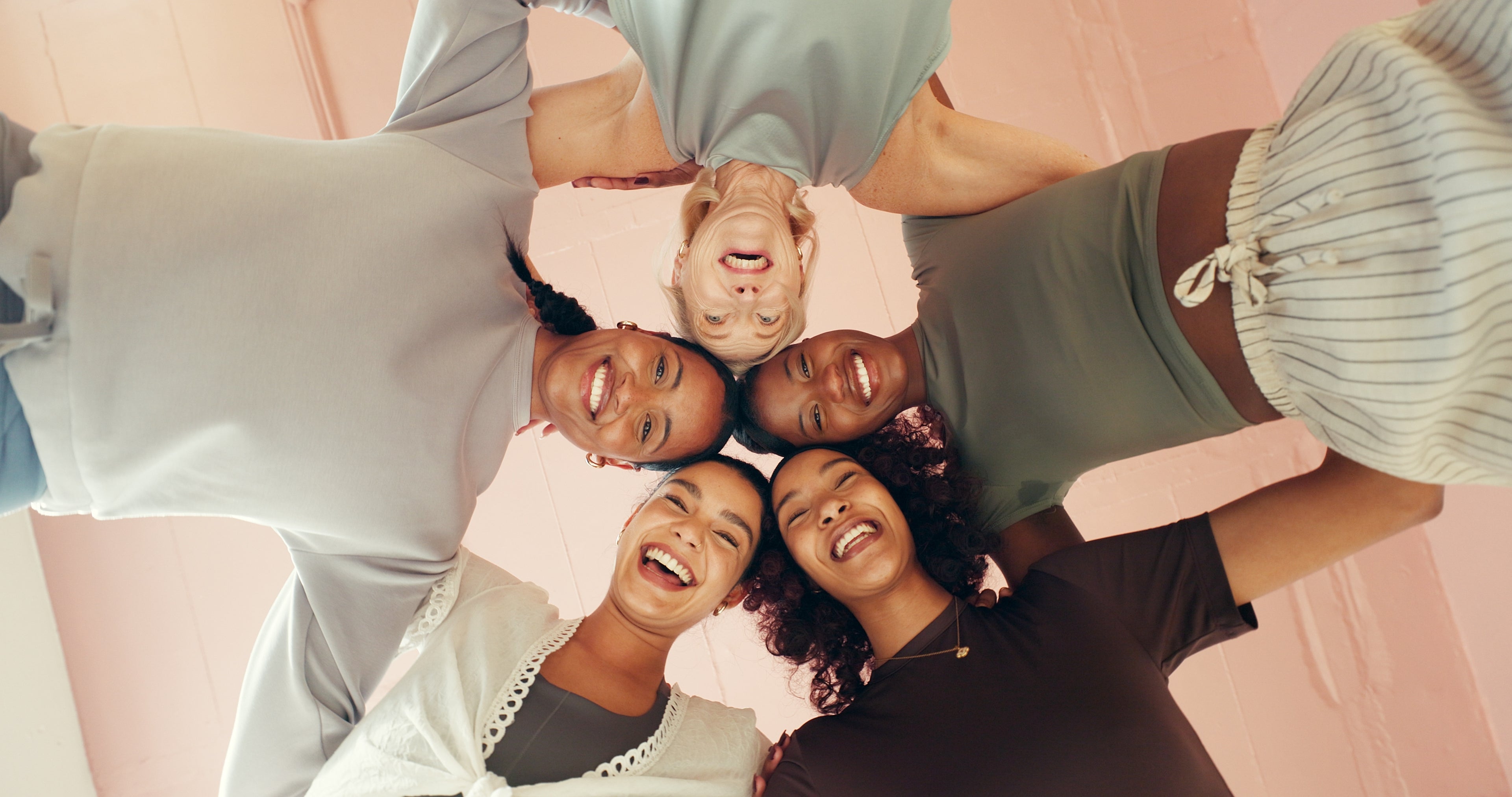 Five people standing in a circle with their heads together against a pink background